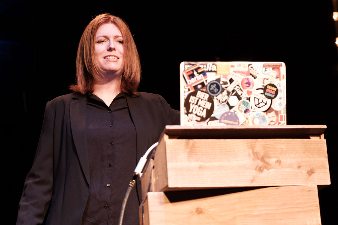 Photo de Charlie Owen lors de sa conférence 'All Constraints are Beautiful'. Elle sourit vers le public, est placée derrière son ordinateur et est habillée en tailleur noir.
